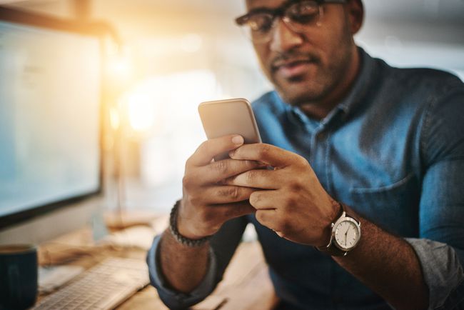 Homem olhando para seu iPhone com um computador em segundo plano