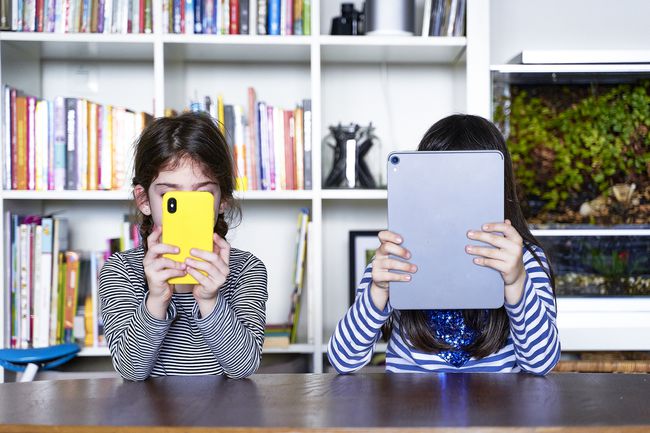 Δύο αδερφές που κάθονται δίπλα δίπλα στο τραπέζι στο σαλόνι χρησιμοποιώντας ηλεκτρονικές συσκευές