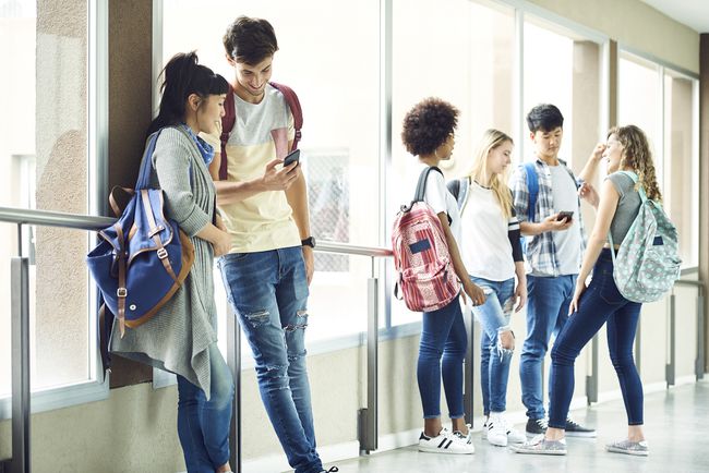 Две группы подростков общаются в школьном коридоре, некоторые используют смартфоны.