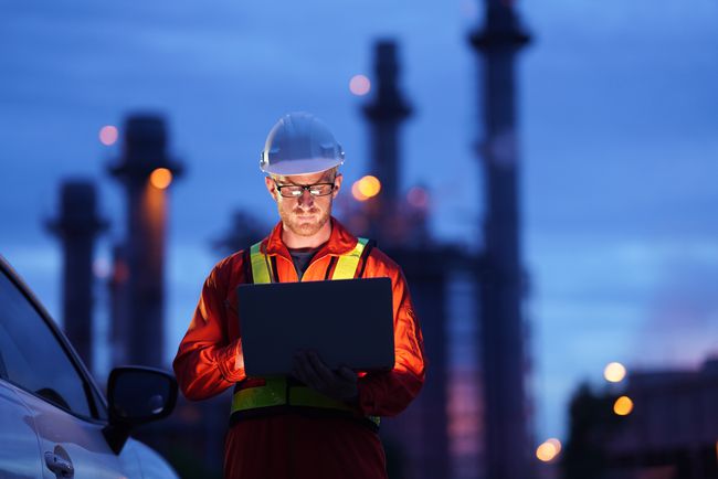 Een bouwvakker met een laptop buiten in de schemering met een fabriek op de achtergrond.