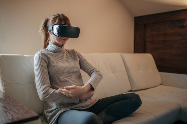 Asmuo, nešiojantis VR ausines sėdėdamas ant sofos ir, atrodo, sūpuoja ant rankų kūdikį.