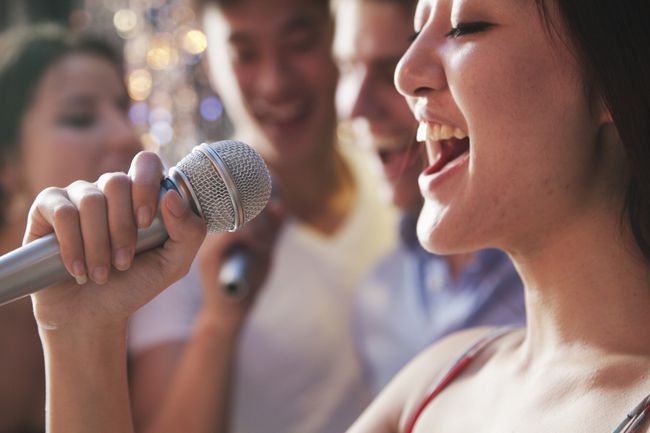 Bližnji posnetek ženske, ki poje v mikrofon s prijatelji v ozadju