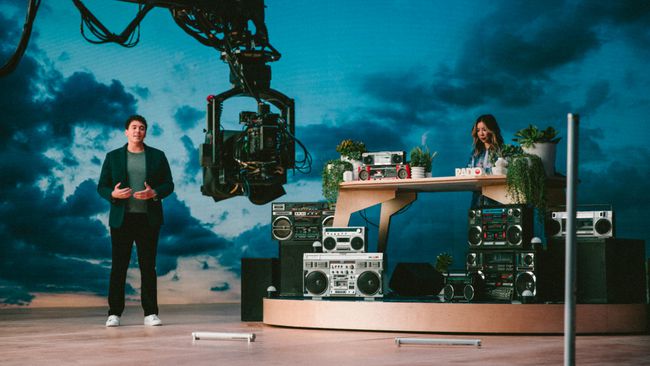 Duas pessoas em um aparelho de TV com aparelhos de som, mesa e fundo de nuvem