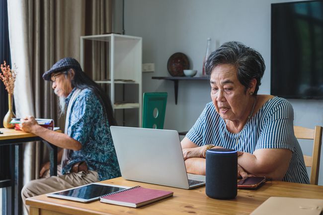 別のシニア大人と一緒に部屋でラップトップで作業しているときにスマートスピーカーに話しかけるシニア大人。