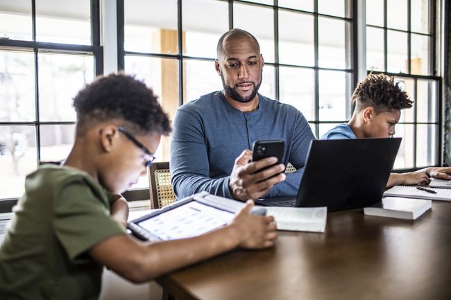 Um adulto usando um smartphone e laptop enquanto duas crianças em idade escolar fazem a lição de casa na mesma mesa.