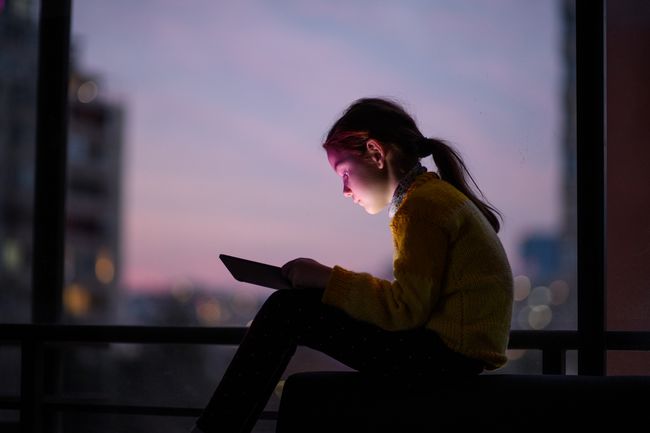 Dijete sjedi ispred prozora u zalasku sunca nad velikim gradom i koncentrira se na tablet računalo.