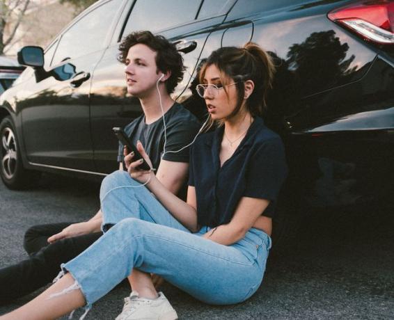 Amigos sentados en el suelo, apoyados en un vehículo, compartiendo música desde un teléfono inteligente con auriculares con cable. 