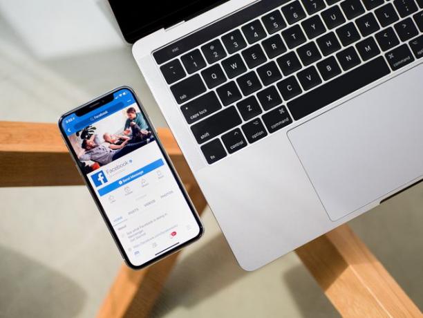 „Facebook“ rodomas išmaniajame telefone, sėdinčiame šalia nešiojamojo kompiuterio ant stiklinio stalo.