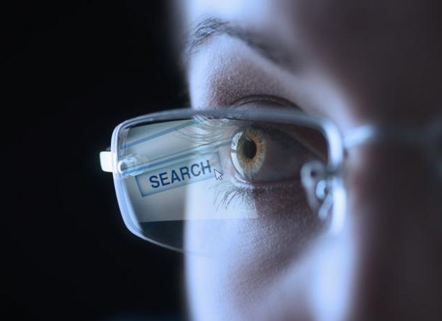 Coseup af et menneskeligt øje, iført briller med en søgeknap, der reflekterer på linsen.