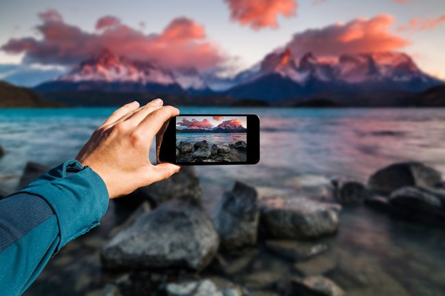 누군가 스마트폰으로 배경화면에 산이 있는 호수의 일몰 사진을 찍고 있습니다.